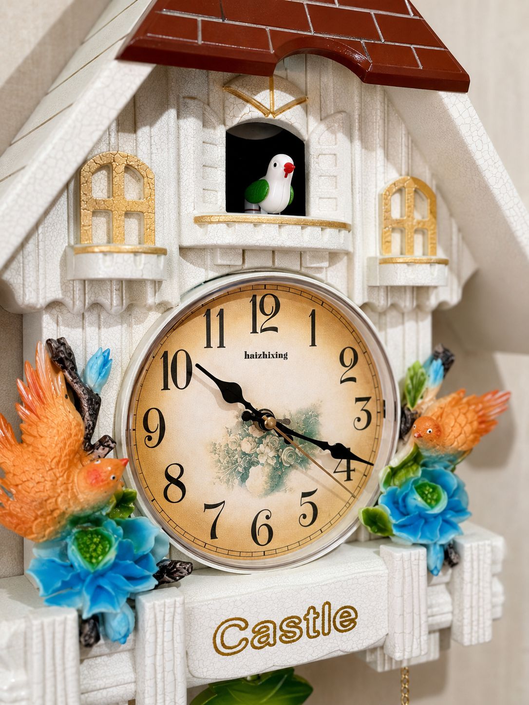 A decorative cuckoo-style wall clock shaped like a white castle with a brown roof, floral accents, and two small birds in windows. The clock face is cream with black numerals and hands, and the word &