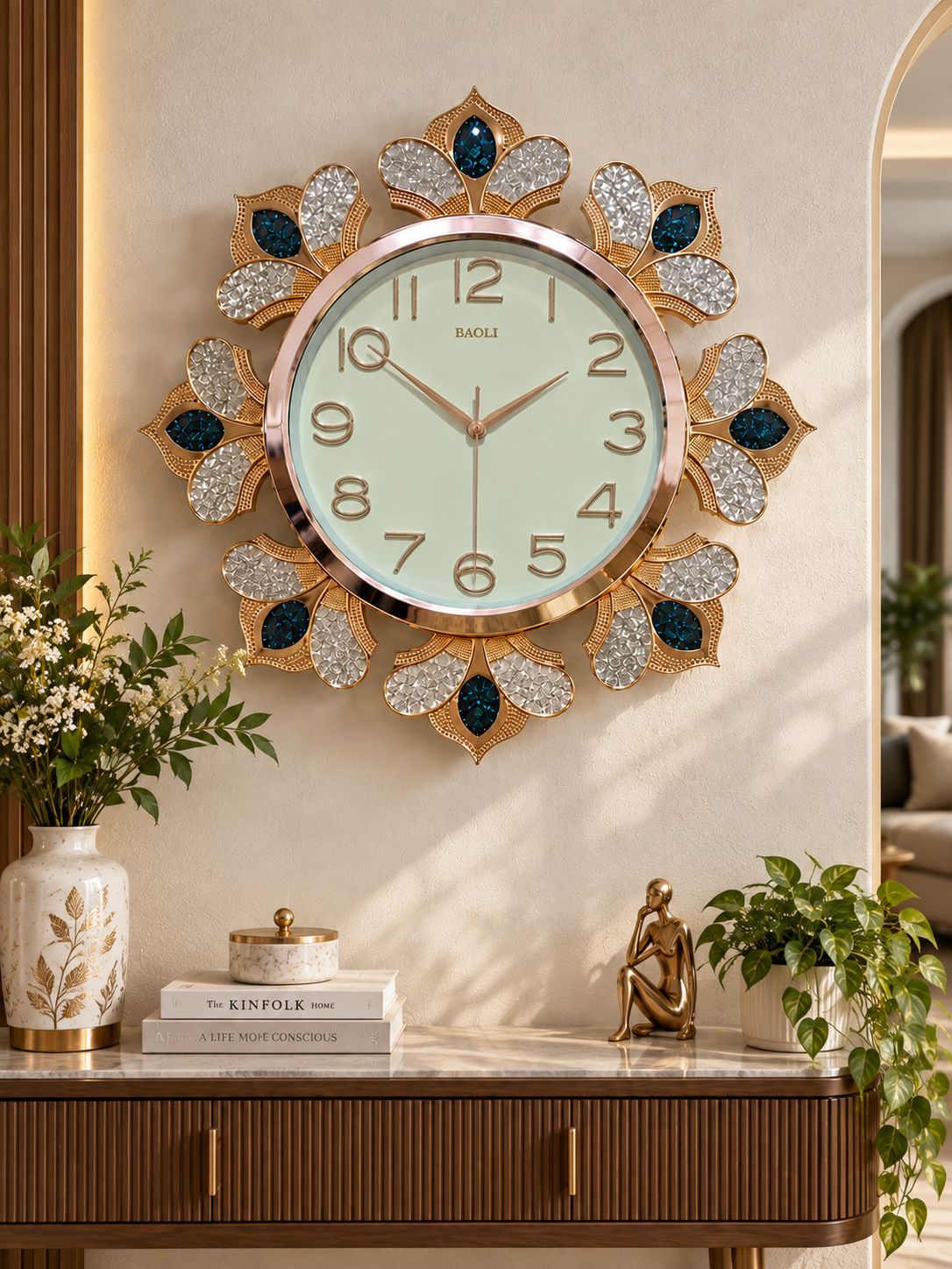 A decorative wall clock with a gold-tone sunburst frame, crystal-embellished petals, and dark blue jewel accents, mounted on a beige wall above a console table in a styled interior.