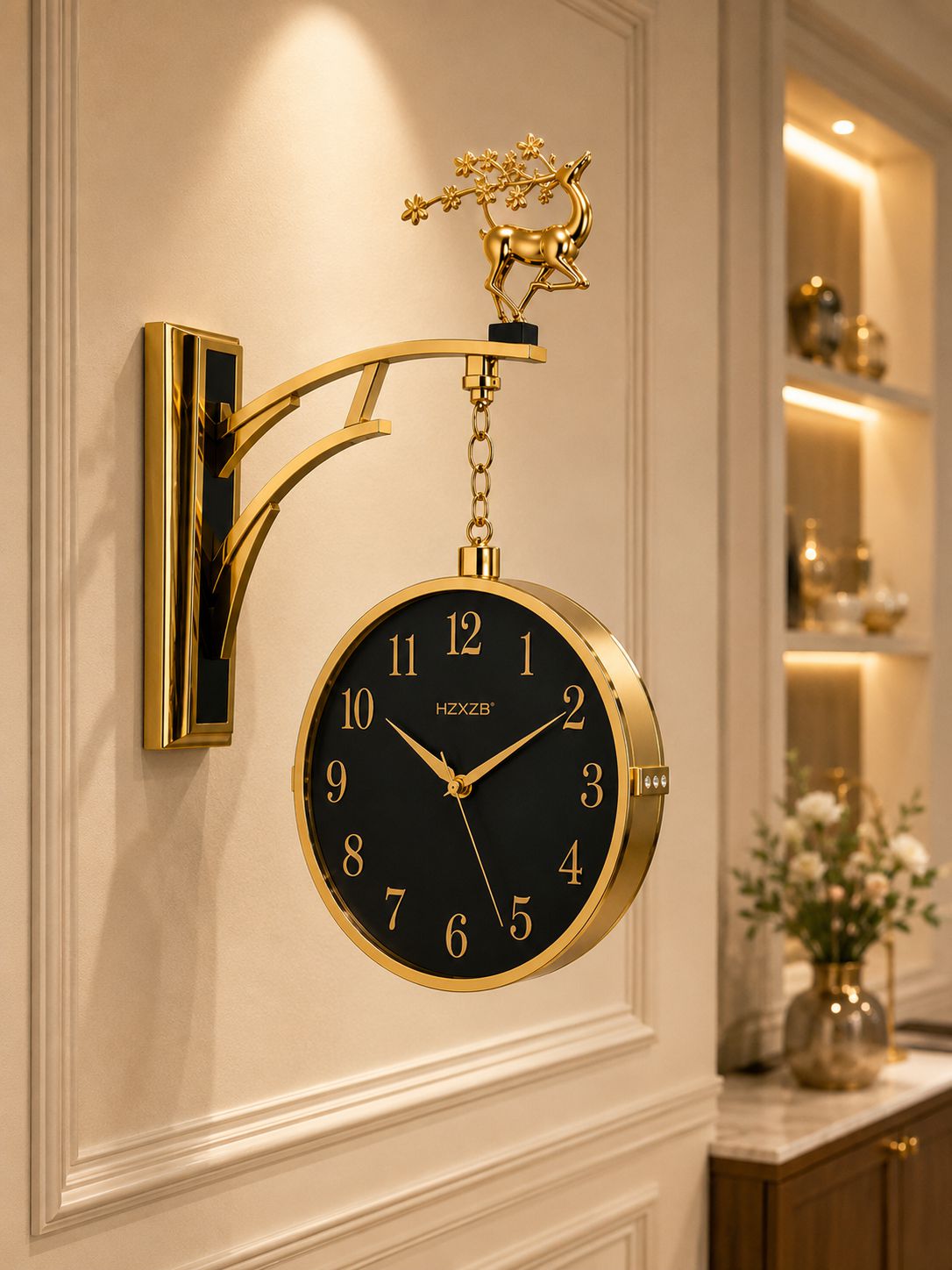 A decorative wall clock with a round black face, gold-tone frame and numerals, hanging from an ornate gold bracket mounted on a cream wall. The bracket features a small deer figurine at the top.