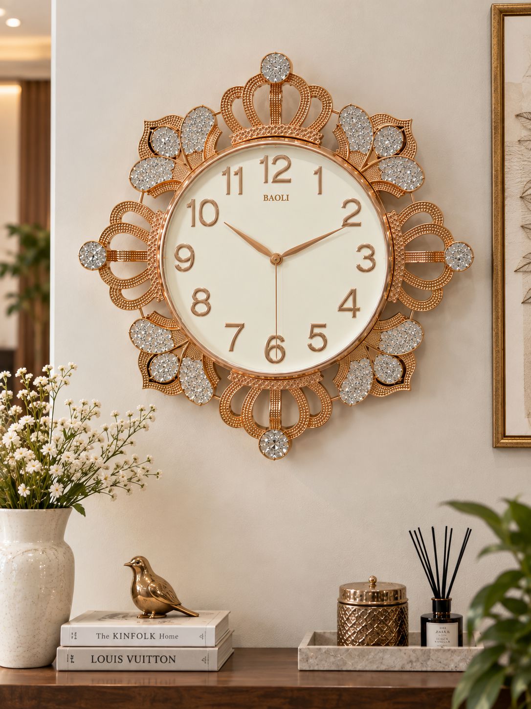 A decorative wall clock with an ornate gold-toned frame, crystal-like rhinestone accents, and a cream dial, mounted on a beige wall above a styled console table with books, flowers, and diffusers.