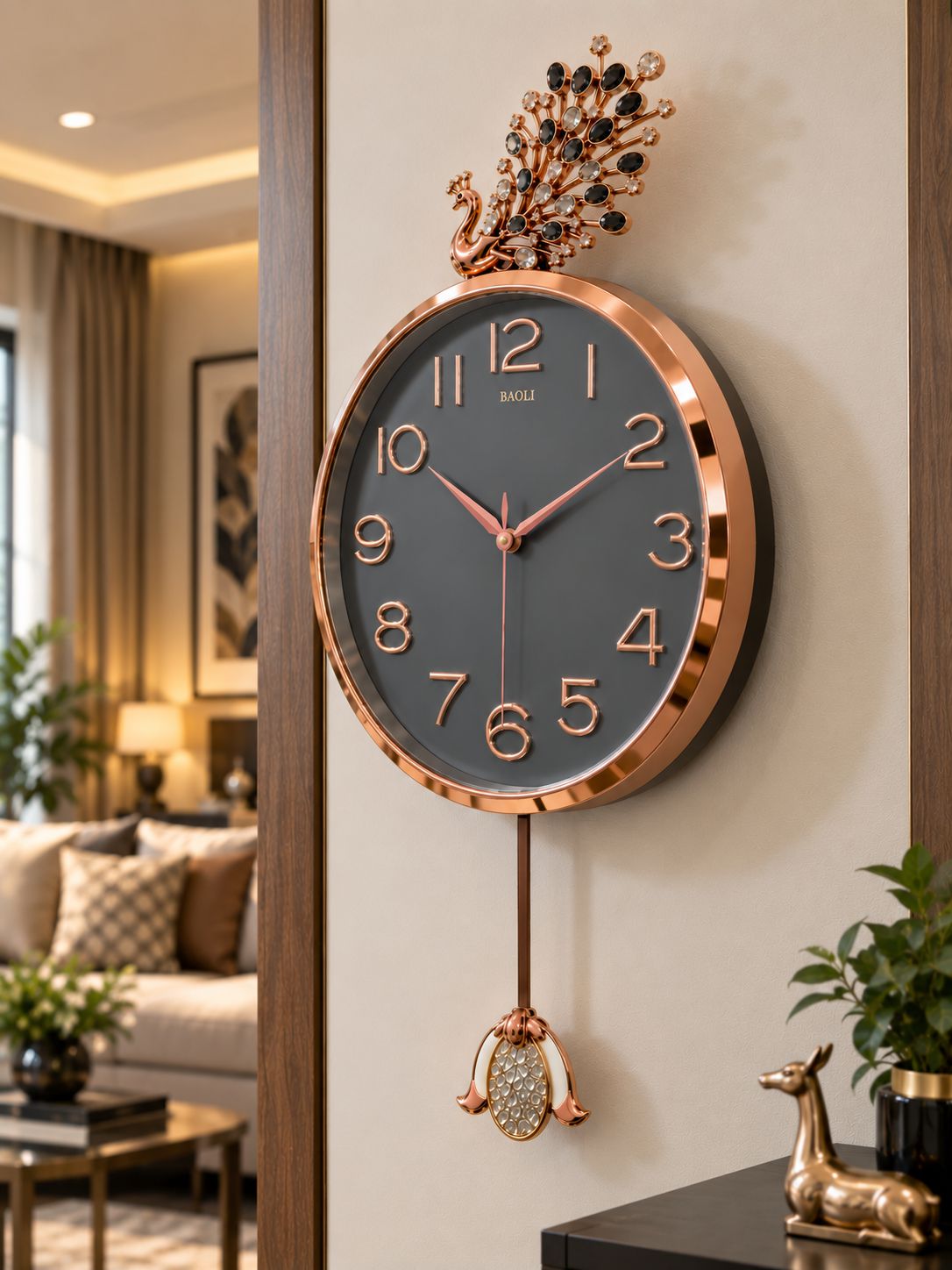 A round wall clock with a dark gray face, rose gold-toned frame and numerals, and a decorative peacock topper with black and clear stones. The clock hangs on a beige wall in a modern living room setting.
