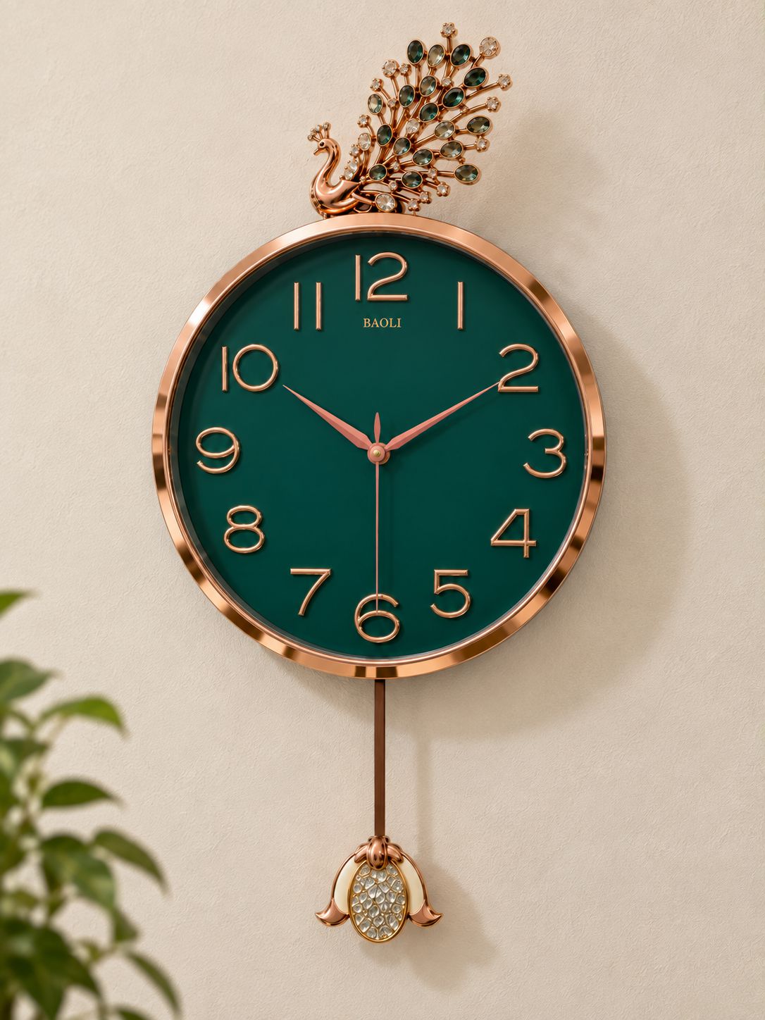 A round wall clock with a dark green dial and rose gold-toned frame, featuring a decorative peacock finial at the top and a dangling ornament at the bottom. The brand name &