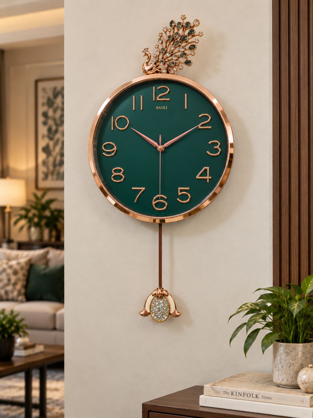 A round wall clock with a dark green face, rose gold-tone frame and hands, a decorative peacock topper, and a hanging ornament, shown mounted on a beige wall in a styled living room.