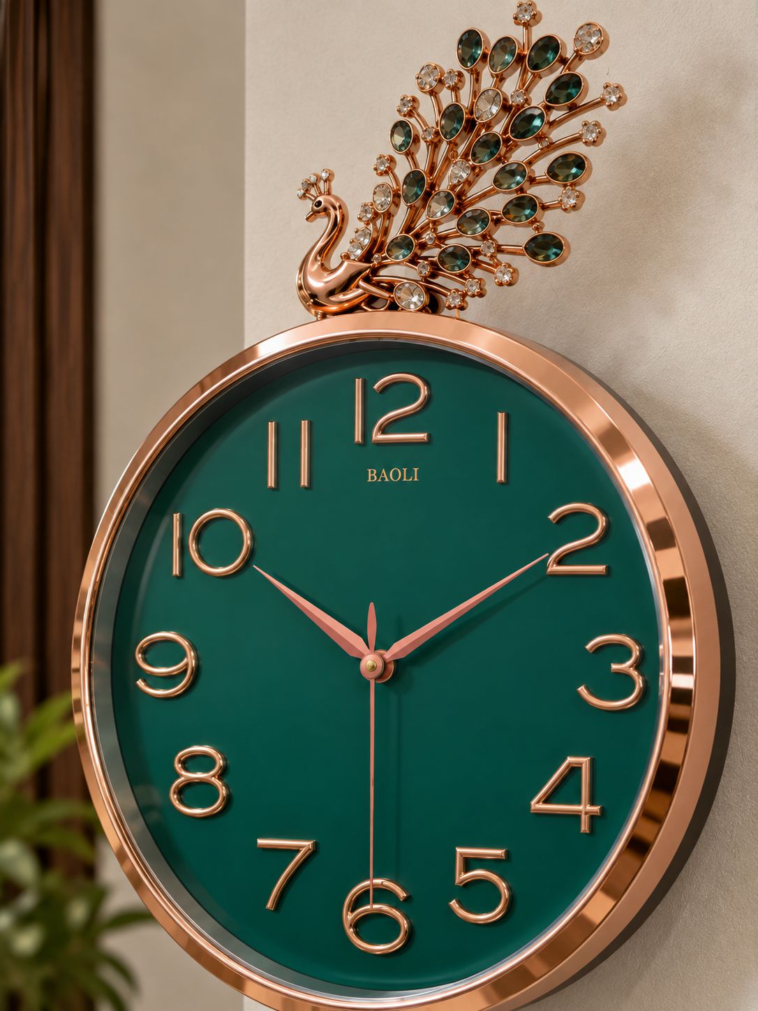 A round wall clock with a rose gold frame and a dark green dial, featuring a decorative peacock topper embellished with green gemstones. The brand name &