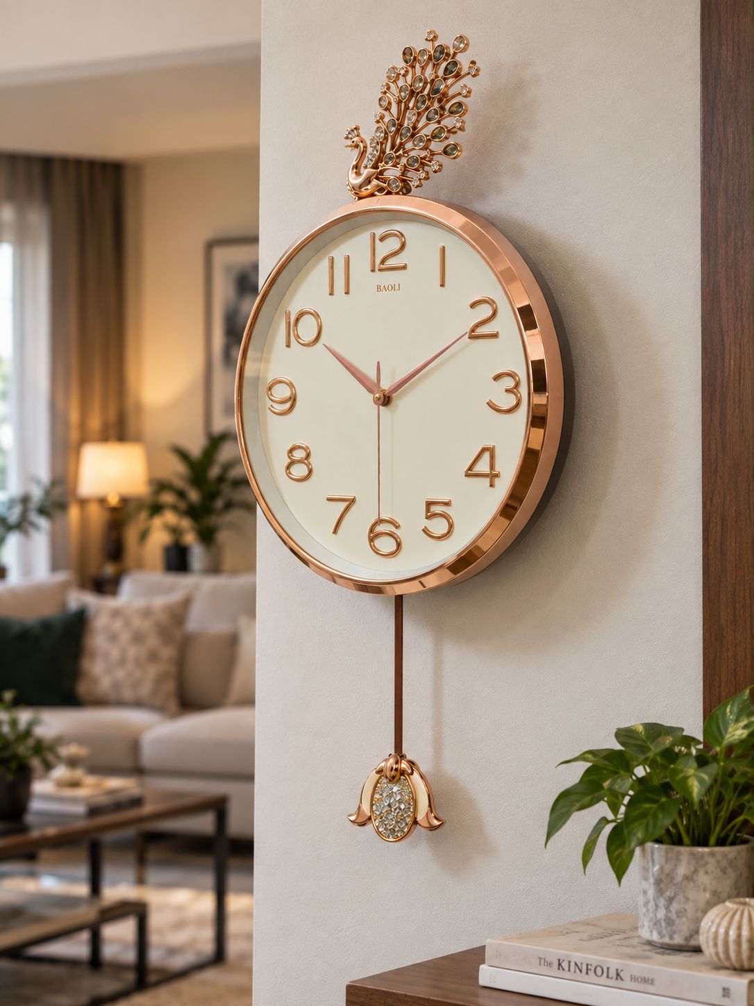 A round wall clock with a rose gold metallic frame, cream-colored face, and matching rose gold hands and numerals, hanging on a beige wall in a modern living room setting.