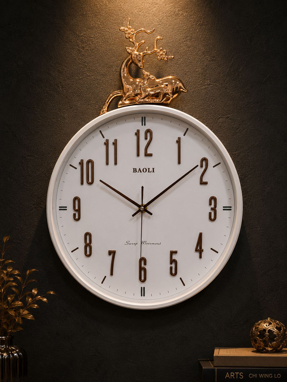 A round wall clock with a white face, black numerals, and black hands, mounted on a dark textured wall. The clock has a decorative gold deer figurine on top and the brand name &
