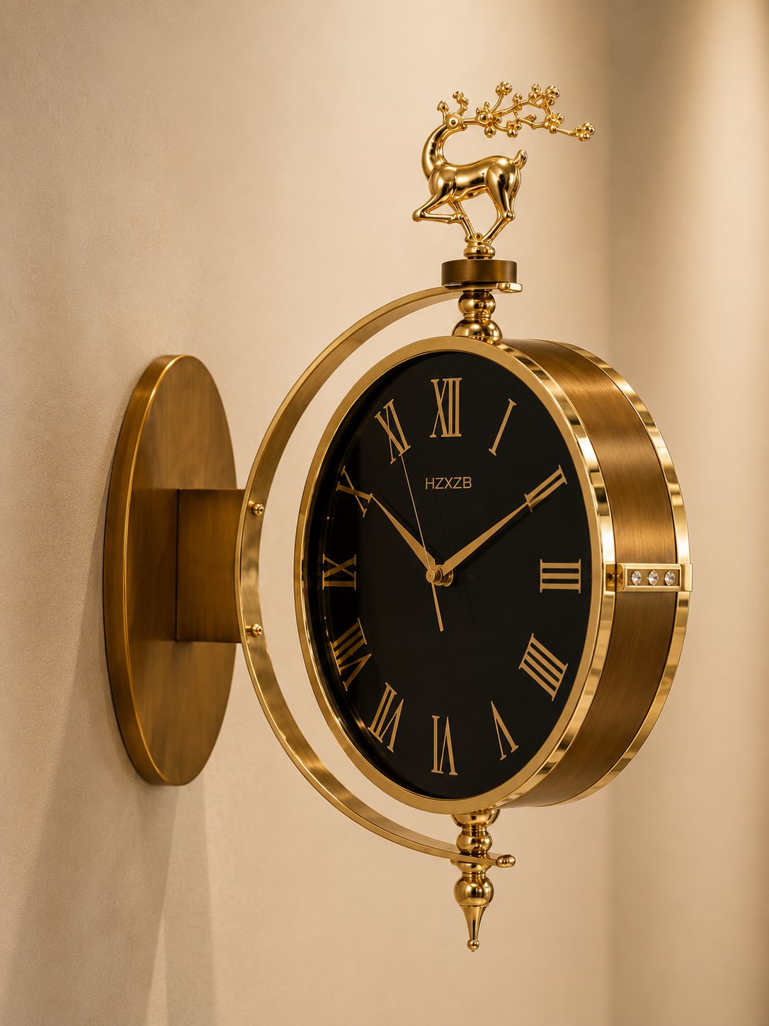 A wall-mounted decorative clock with a black dial, gold Roman numerals, and a polished gold metal frame and wall mount. A small gold deer figurine sits atop the frame, and the brand name &