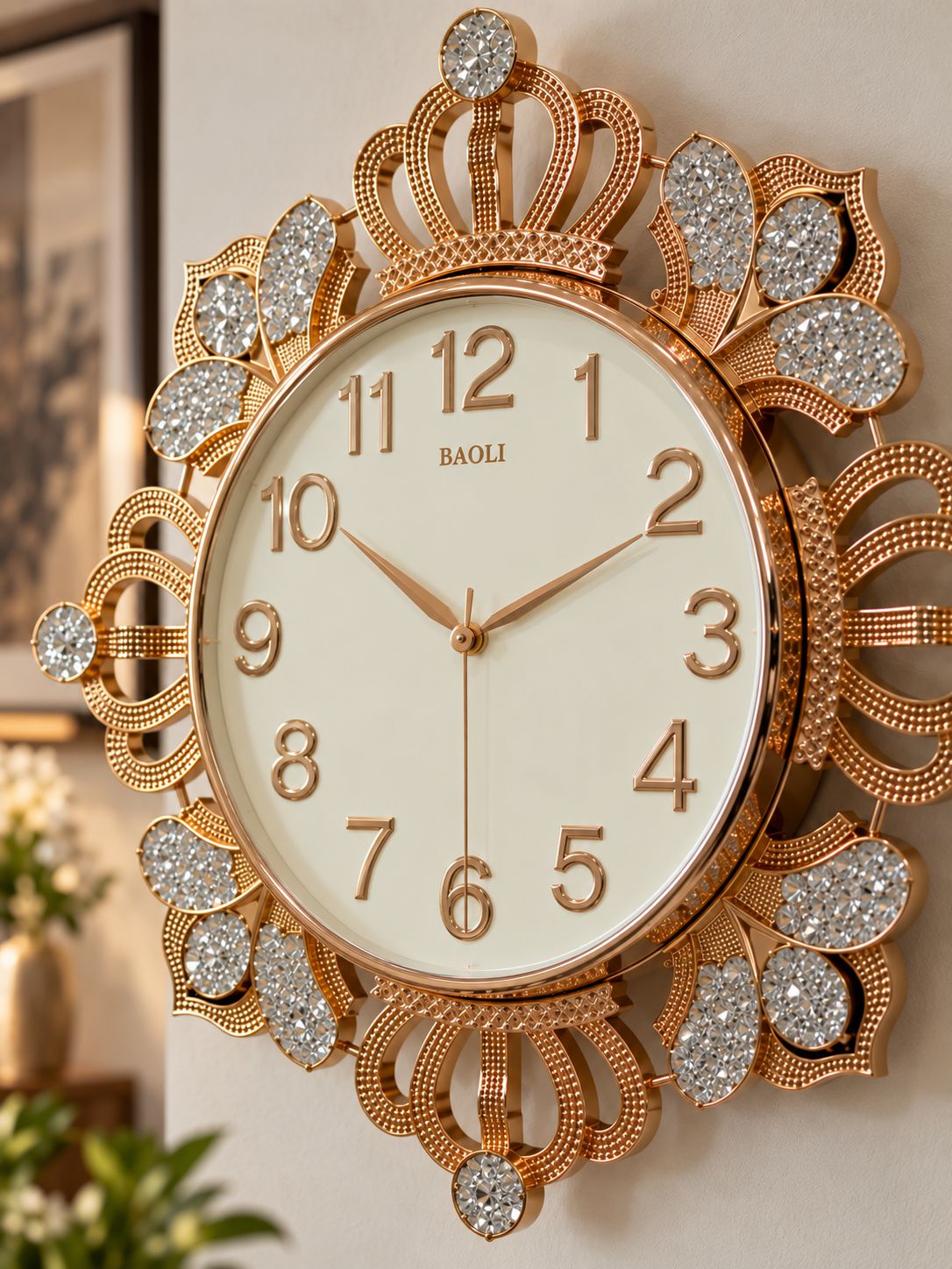 An ornate Baoli wall clock with a gold-tone filigree frame, crystal-embellished details, and a cream-colored dial, shown mounted on a light beige wall.