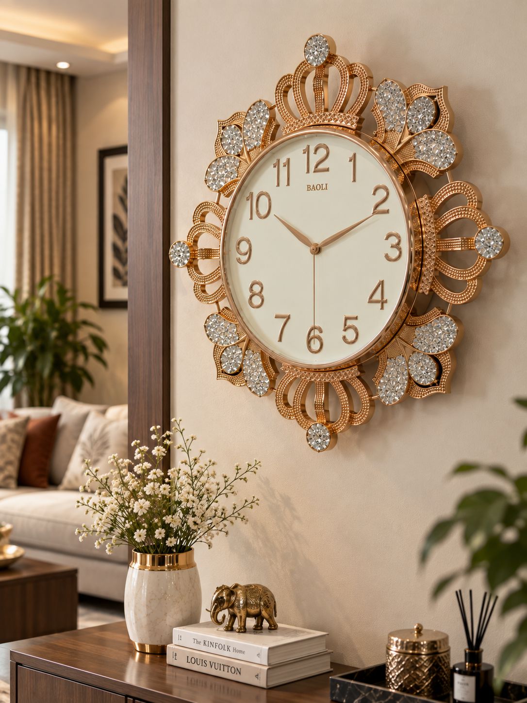 An ornate gold wall clock with a cream-colored face and crystal-like embellishments, mounted on a beige wall in a styled living room setting.
