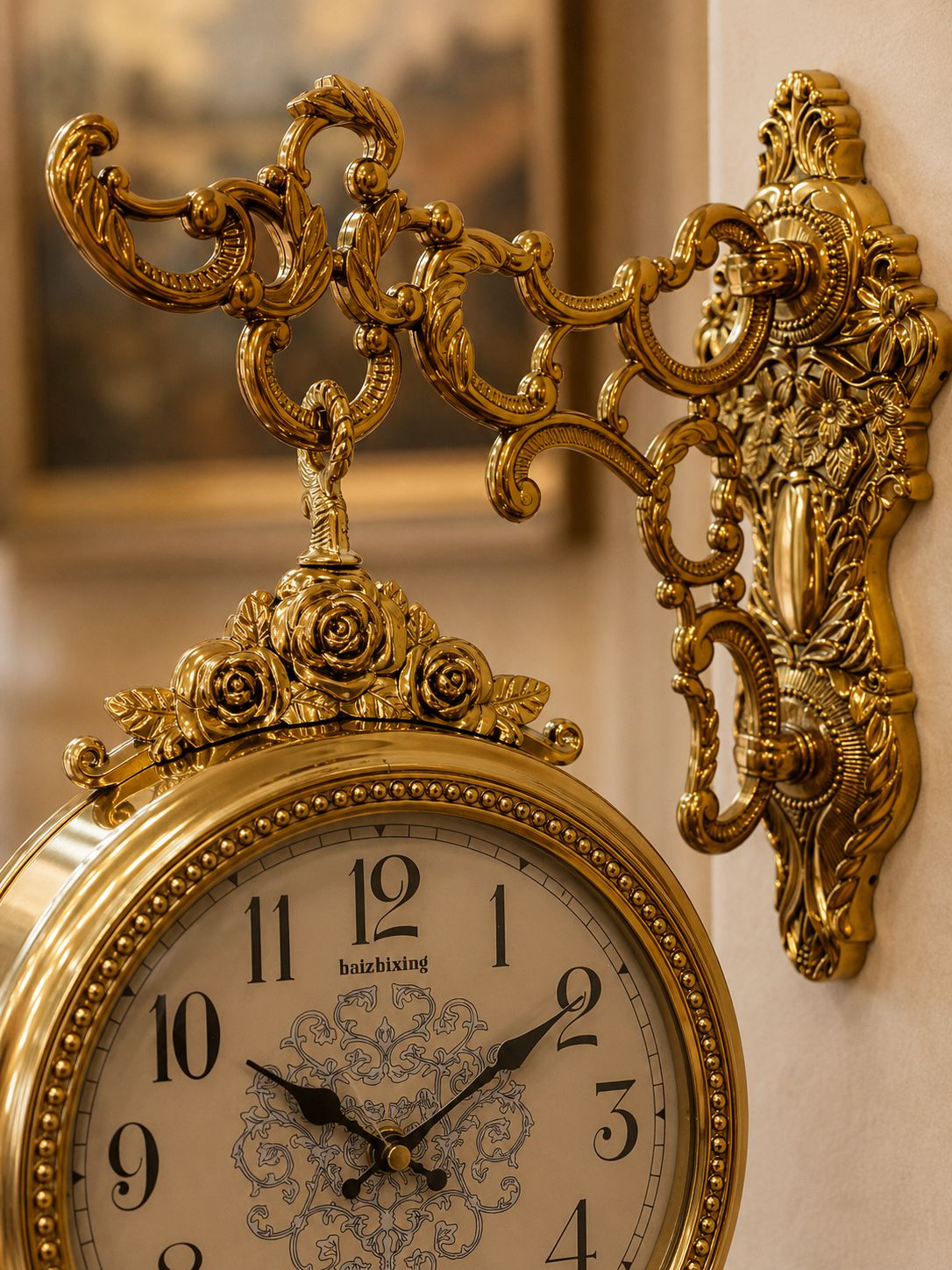 An ornate gold wall clock with a decorative scroll bracket and cream-colored dial, mounted on a beige wall.