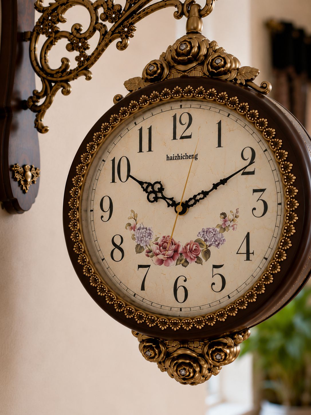 An ornate vintage-style wall clock with a dark brown frame, gold-tone metal scrollwork mount, and a cream dial decorated with black Arabic numerals, floral artwork, and the brand name &