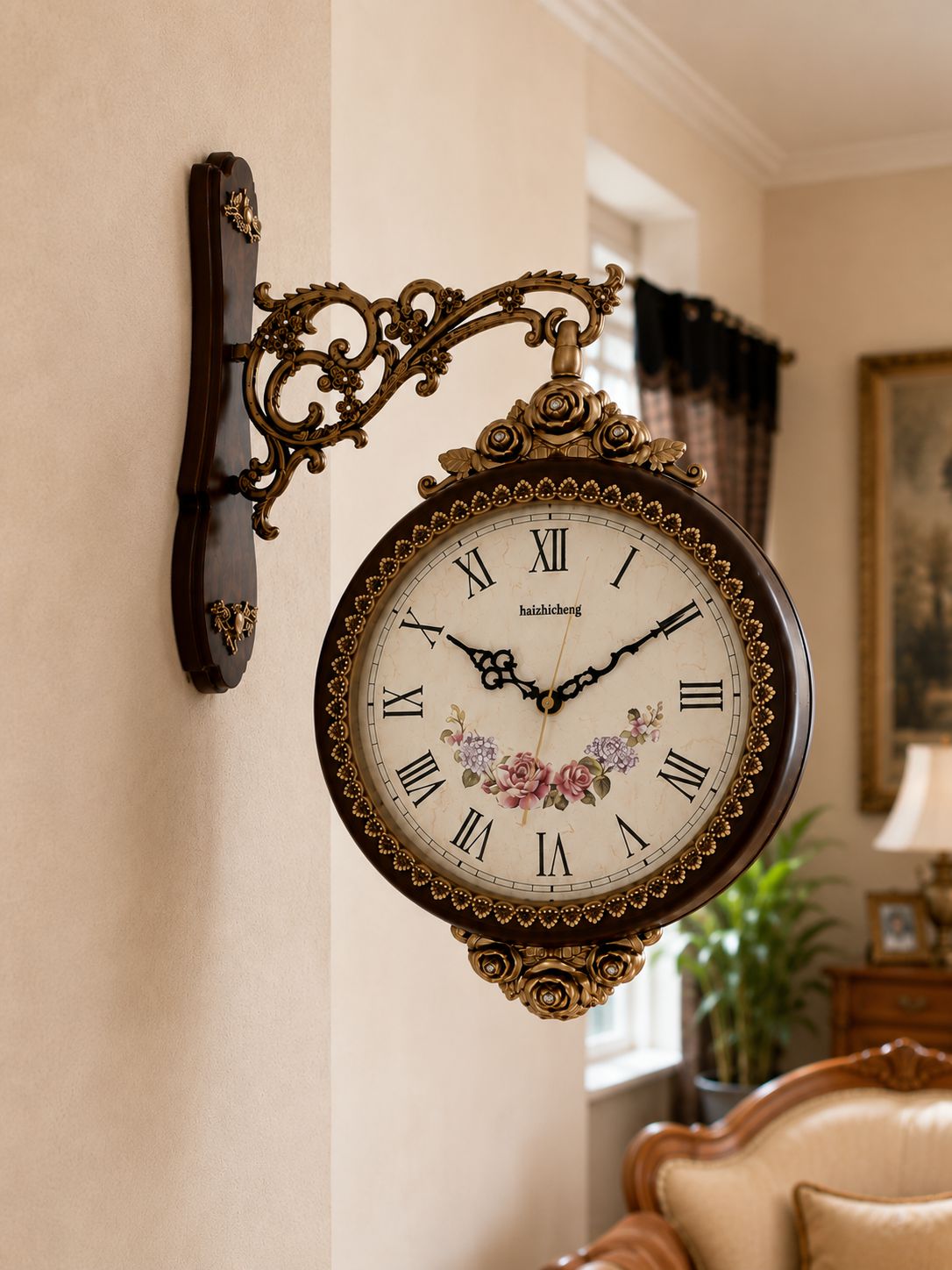 An ornate wall-mounted clock with a dark brown and gold metal bracket, Roman numerals, and floral details on the clock face, shown hanging on a beige wall in a decorated room.