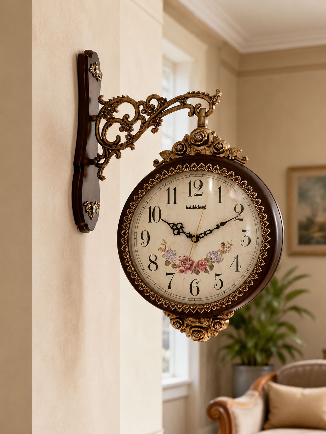 An ornate wall-mounted clock with a dark brown wood frame, gold-toned scrollwork bracket, and floral clock face, shown hanging on a beige wall in a styled interior room.