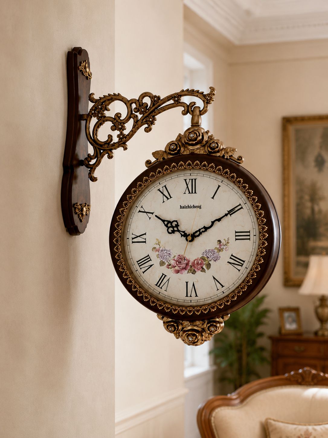 An ornate wall-mounted clock with a dark brown wood frame, gold-toned scrollwork bracket, white dial, black Roman numerals, and floral decoration near the bottom, shown hanging on a beige wall in a styled interior room.