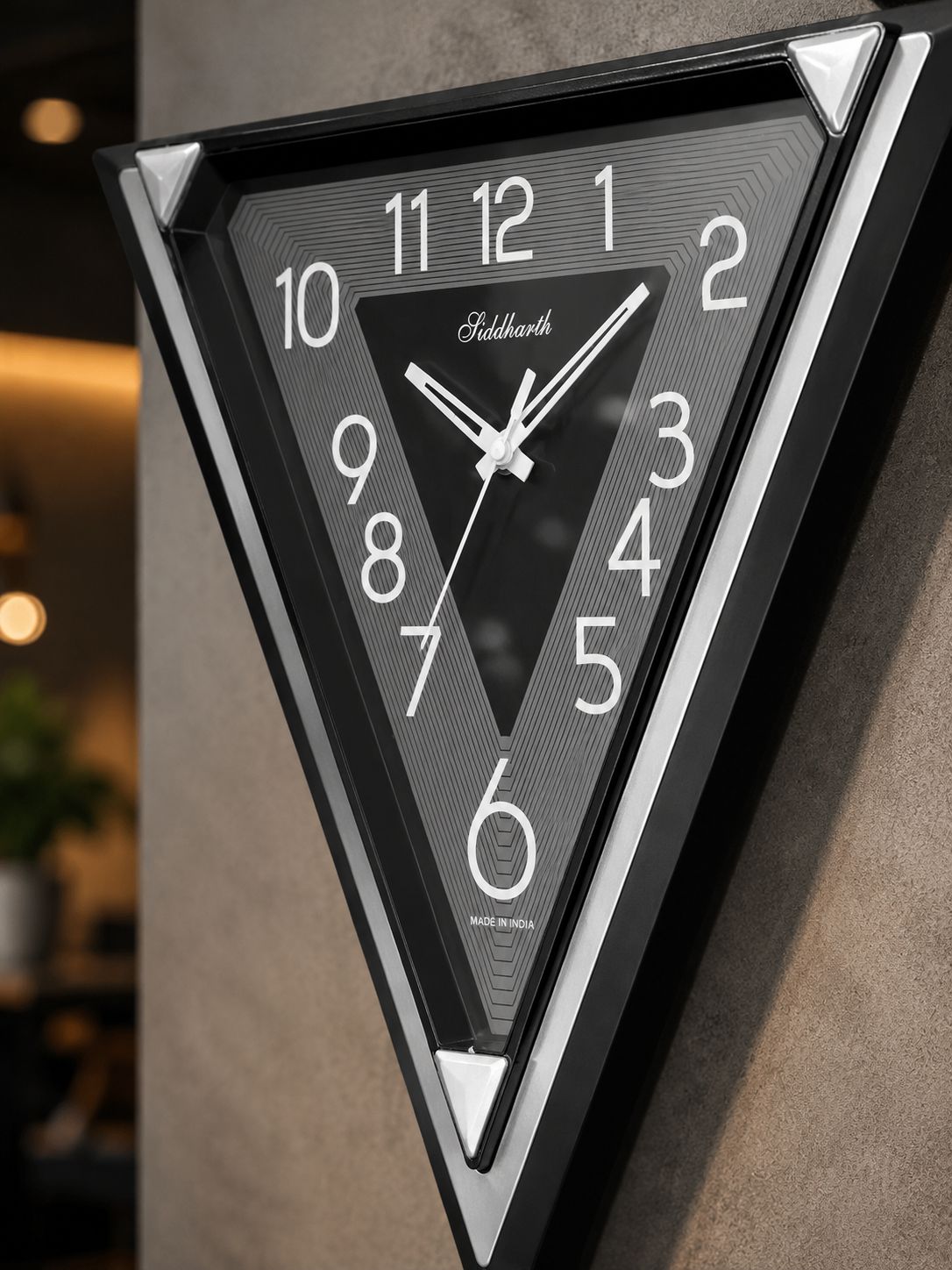 A triangular wall clock with a black frame and textured dark gray face, featuring white Arabic numerals and hands. The brand name &