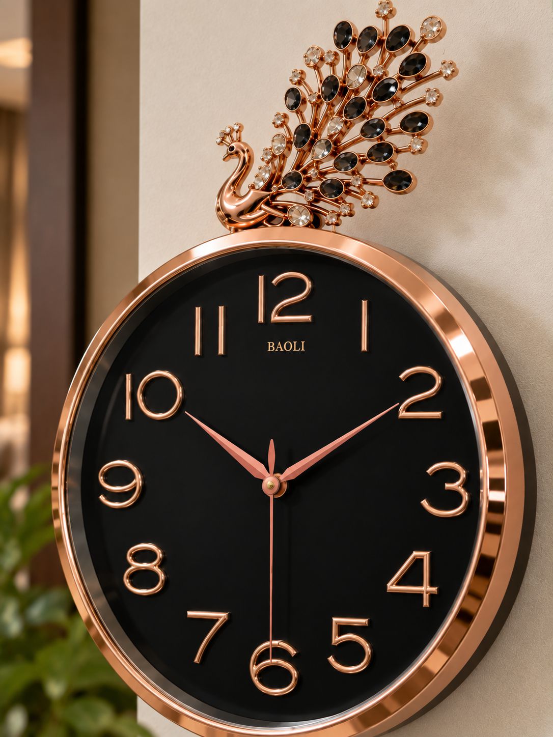 Round wall clock with a black face, rose gold-tone frame and numerals, and a decorative peacock topper with black gemstone-like accents, shown mounted against a beige wall.