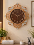 A decorative wall clock with a gold-toned ornate frame, crystal-like embellishments, and a dark brown clock face, mounted on a beige wall above a styled tabletop with books, a plant, and flowers.