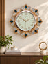 A decorative wall clock with a gold-toned sunburst frame, silver glitter petal details, and dark blue jewel accents, mounted on a beige wall above a wooden console table with books and plants.