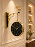A decorative wall clock with a round black face, gold-tone frame and numerals, hanging from an ornate gold bracket mounted on a cream wall. The bracket features a small deer figurine at the top.