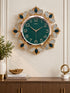 A decorative wall clock with a round green face and ornate gold-tone frame, surrounded by butterfly-like motifs with dark blue and silver rhinestone details. The clock is mounted on a cream wall above a wooden console table with books and decor.