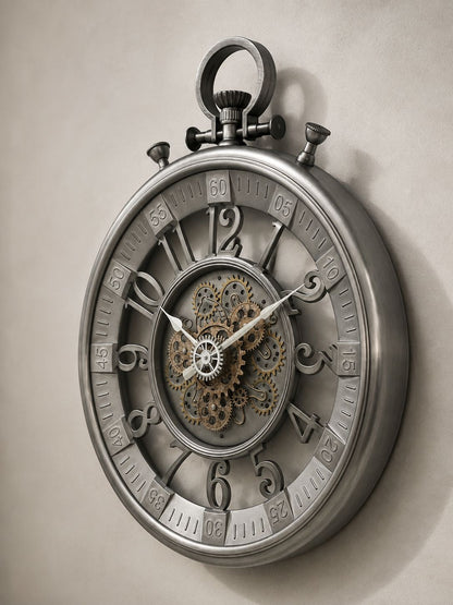 A large decorative wall clock designed like a pocket watch, featuring a metallic silver outer casing, exposed internal gears and cogs at the center, and raised Arabic numerals around the dial, shown against a plain light beige wall.