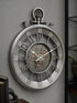 A large round wall clock styled like a pocket watch, with a metallic silver outer frame, exposed inner gears, and ornate black Arabic numerals, mounted on a gray wall.