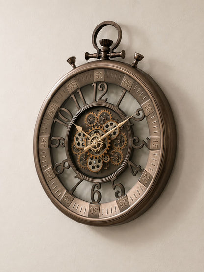 A large round wall clock styled like a vintage pocket watch, featuring a bronze-toned metal frame, exposed internal gears, and ornate Arabic numerals, mounted on a plain beige wall.