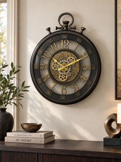 A large round wall clock with a dark metal frame and exposed inner gears, mounted on a beige wall above a wooden console table with books and decor.