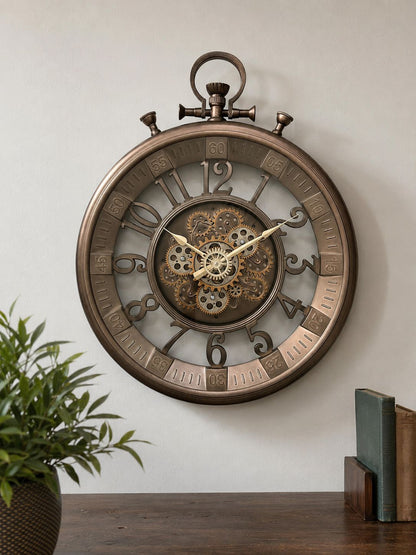 A large round wall clock with a vintage stopwatch design, featuring a bronze-toned metal frame, exposed internal gears, and large Arabic numerals, mounted on a light wall above a wooden surface with a plant and books.