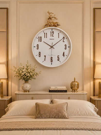 A large round wall clock with a white face and black Arabic numerals hangs above a bed in a warm, beige-toned bedroom. The clock has a decorative gold horse figure at the top and the brand name &