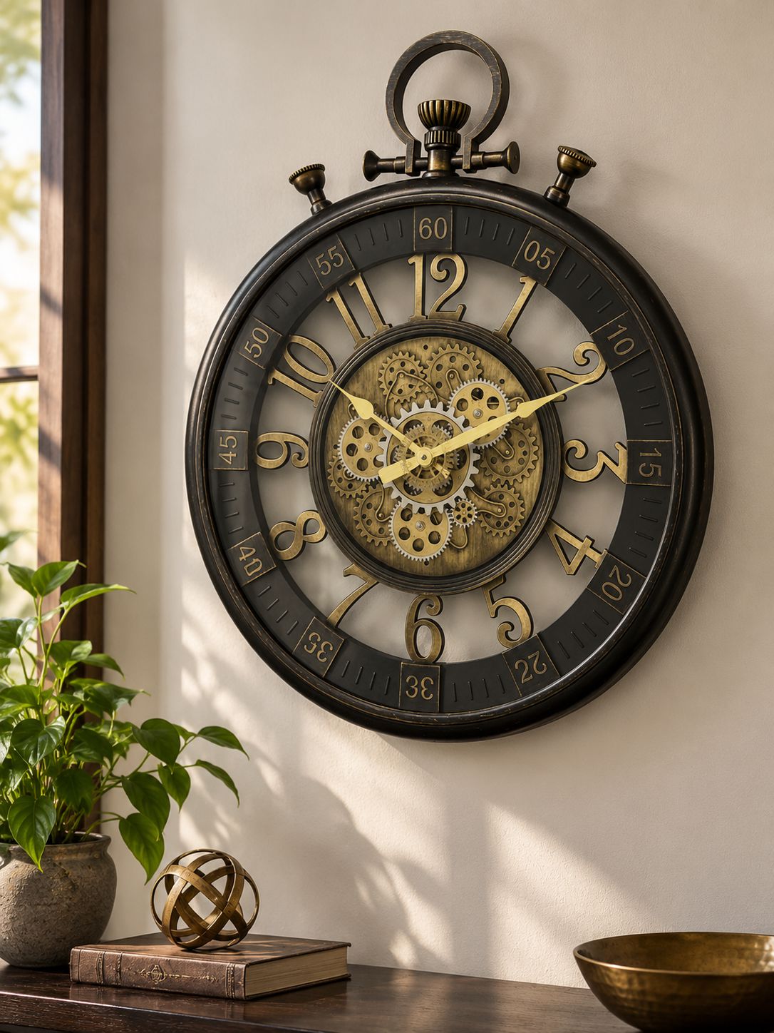 A large vintage-style wall clock with a dark round frame, gold numerals, and a visible gear-like center, mounted on a beige wall above a wooden table with a plant and decorative objects.