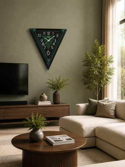 A modern triangular wall clock with a dark green and black frame and white numerals, hanging on a sage green wall in a styled living room with a beige sectional sofa, wooden coffee table, and potted plants.