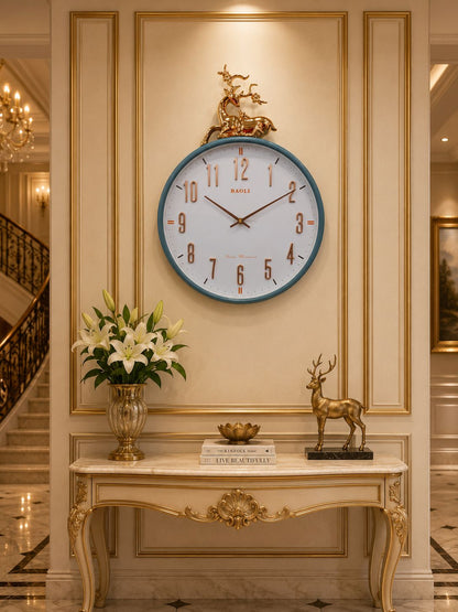 A round wall clock with a blue rim and gold decorative horse-and-rider sculpture above it, mounted on a cream paneled wall above a console table with flowers and a deer statue.