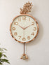 A round wall clock with a rose gold metallic frame and a cream-colored dial, featuring a decorative peacock topper and a dangling peacock weight with a rhinestone-encrusted center, mounted on a light beige wall.