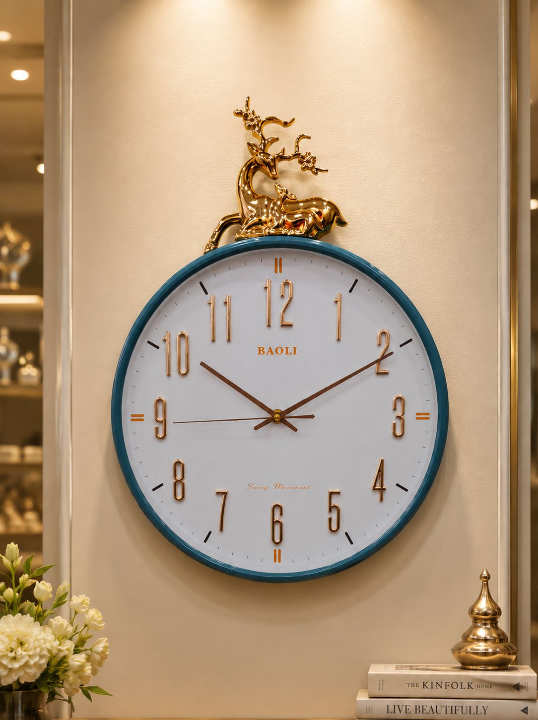 A round wall clock with a white face, blue rim, and gold-tone numerals and hands, mounted on a beige wall. A gold deer sculpture sits above the clock.