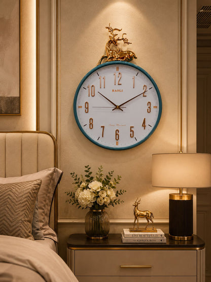 A round wall clock with a white face, gold Arabic numerals, and a teal-blue outer frame, mounted above a bedside table in a warmly lit bedroom. The clock has a decorative gold deer sculpture on top.