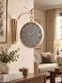 A wall-mounted clock with a round gray face, gold Roman numerals, and a gold curved bracket, shown hanging in a styled living room interior.