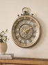 An antique-style wall clock shaped like a pocket watch hangs on a beige wall above a wooden console table. The clock features a distressed bronze finish, skeletonized dial showing intricate inner gears, and raised Arabic numerals.
