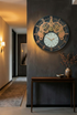 Decorative wall clock with mechanical design on a dark wall in a room with a table and lamp.