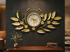 Large gold metal wall clock with leaf design on grey wall for living room decor.