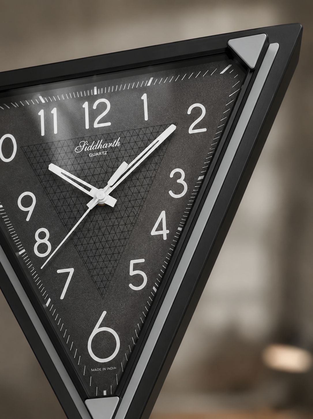 A close-up of a triangular wall clock with a black frame and textured dark gray dial, featuring white Arabic numerals and hands. The brand name &