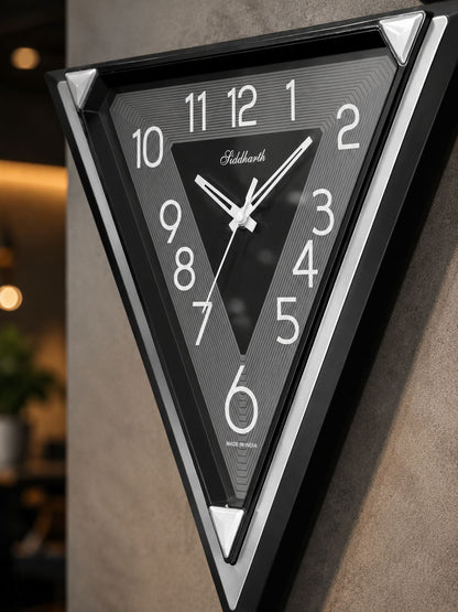 A triangular wall clock with a black frame and textured dark gray face, featuring white Arabic numerals and hands. The brand name &