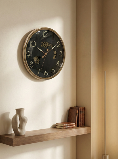 Wall clock on a beige wall with a shelf below containing books and a vase.